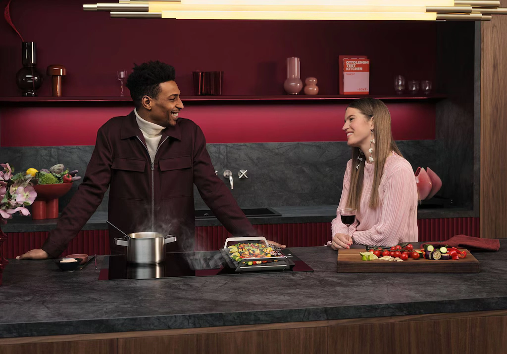 Twee personen koken samen aan een kookeiland met AEG inductiekookplaat en geïntegreerde afzuiging in een moderne keuken met rode accenten